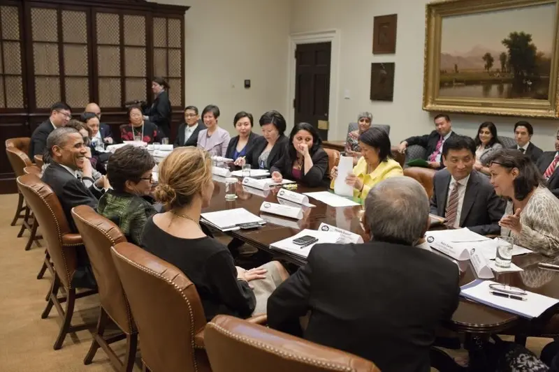 A crowd of people around a long conference table. Danner leans forward, speaking, and President Obama looks at her, smiling.