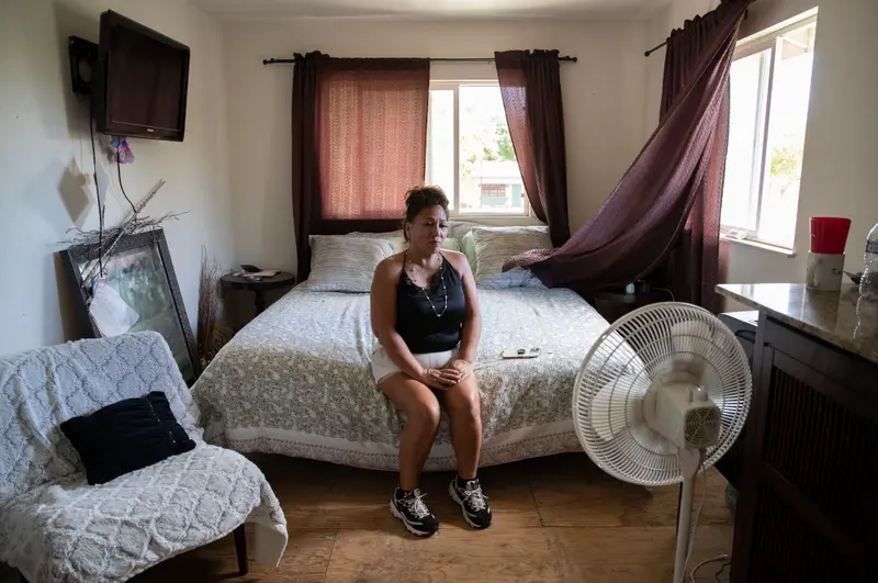 A woman sits on a bed in a room with a bare plywood floor.